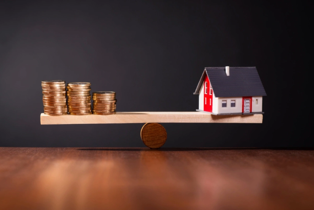 balance avec à gauche des pièces et à droite une maquette de maison pour symboliser la gestion de patrimoine